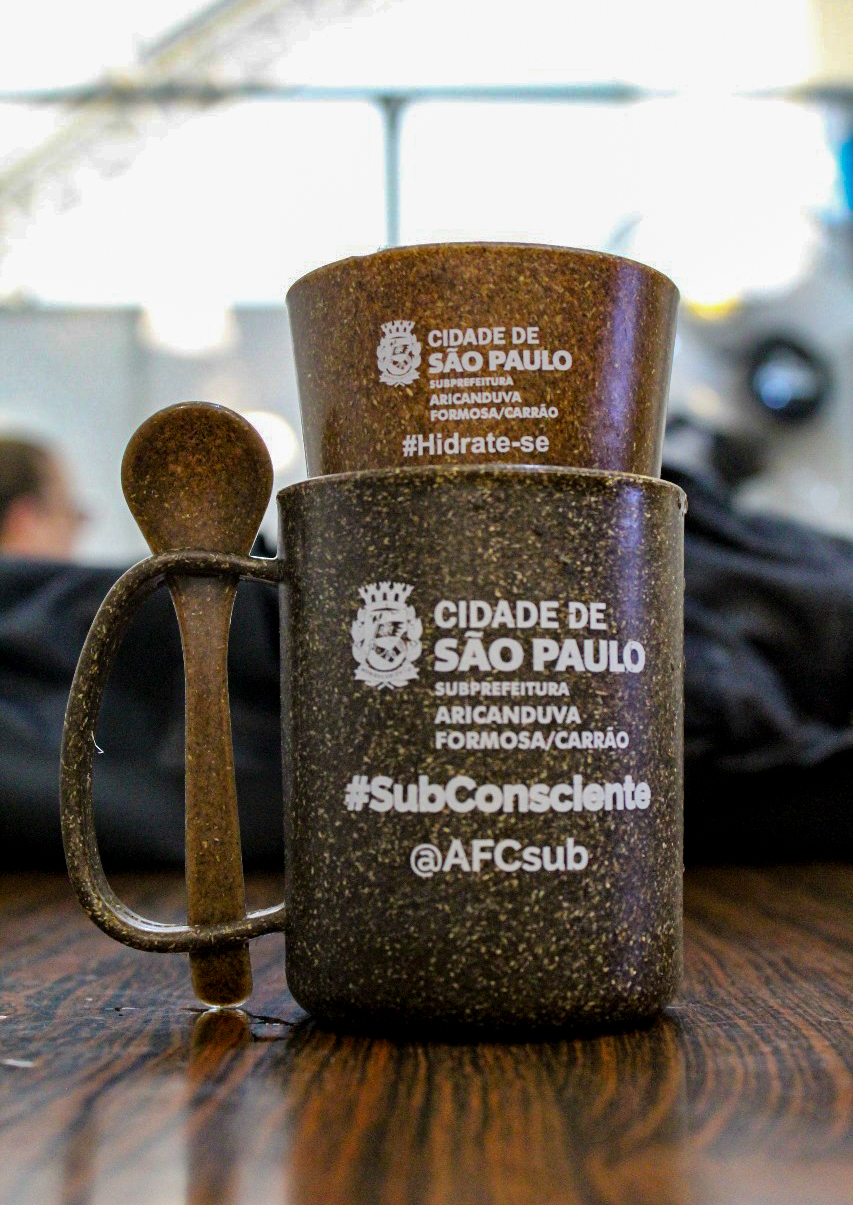 A imagem mostra a foto da caneca e do copo ecológico feitos com material de fibra de coco. Na caneca, visualiza-se o logo da Sub AFC, embaixo os dizeres: #SubConsciente, e, por fim, o usuário da Sub nas redes sociais ("@AFCsub"). O copo está dentro da caneca e ambos estão postos em cima de uma mesa.